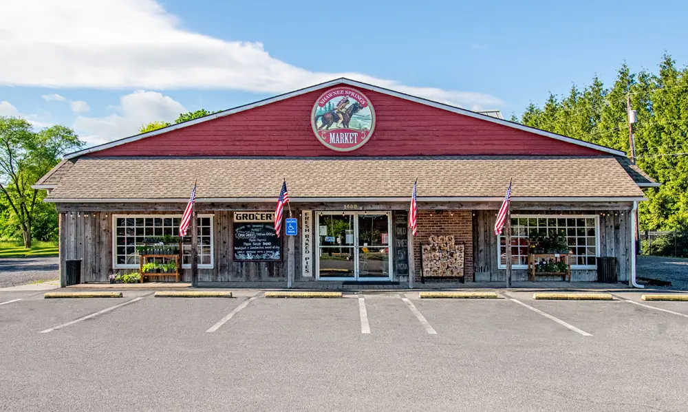 A rustic market building with American flags, parking lot, and greenery. Prominent sign on red facade. Bright, clear day.