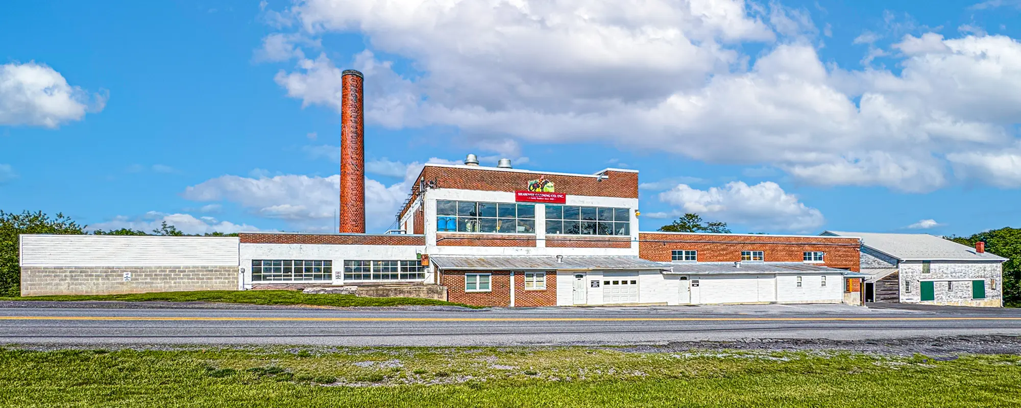 A large, vintage brick building with a smokestack, surrounded by open grass, under a partly cloudy sky. Factory-like structure with a retro sign.