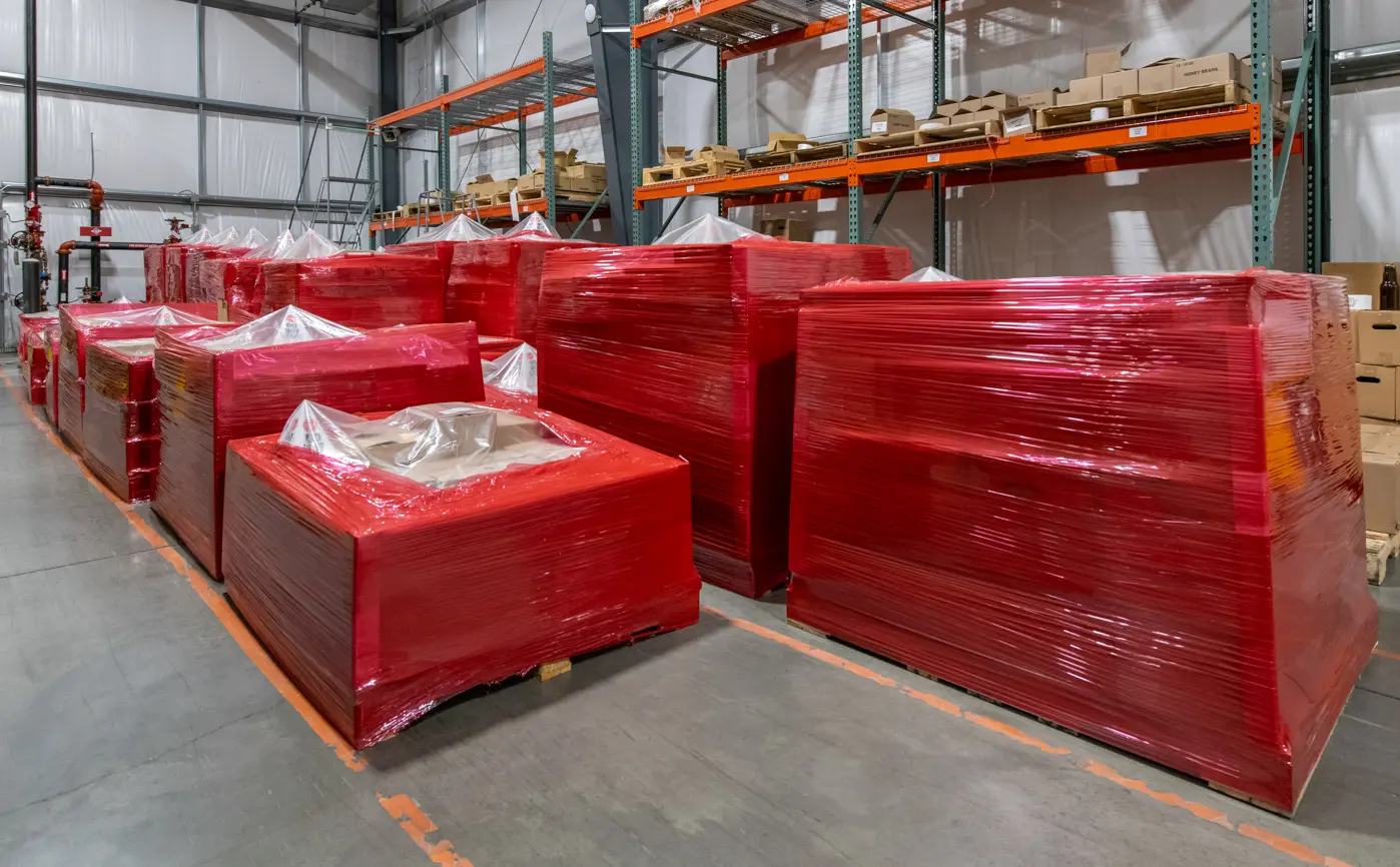 Several large items wrapped in red plastic are stacked in a warehouse. Metal shelves hold various boxes in the background.