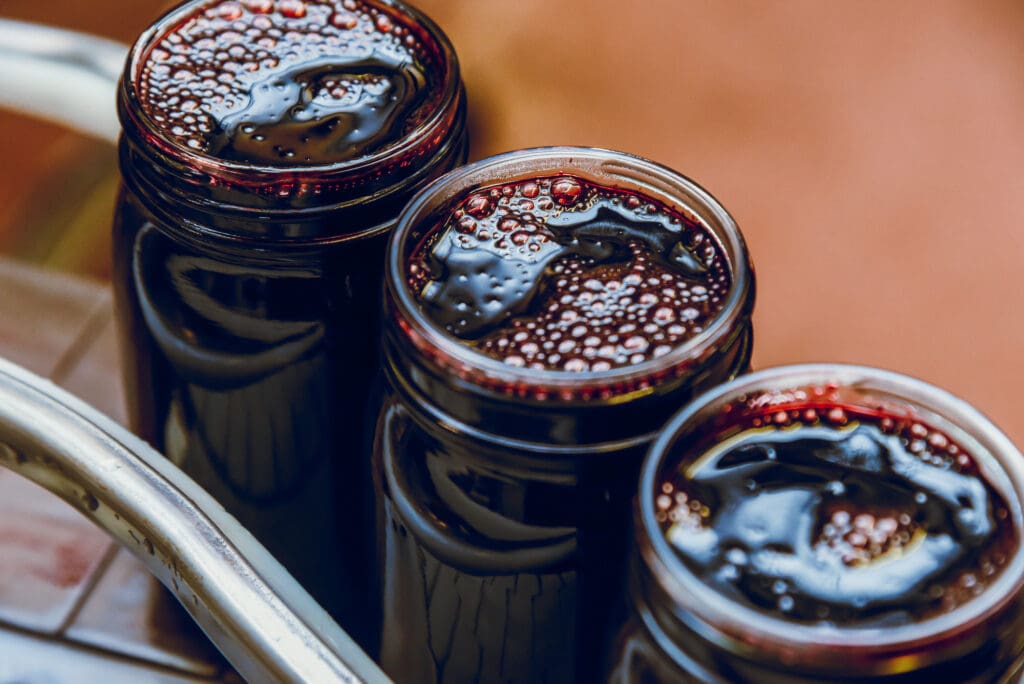Three jars filled with dark red jam, tightly sealed and placed on a table. The surface appears glossy, with bubbles on top.