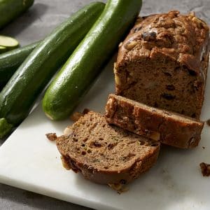 A loaf of zucchini bread with sliced pieces is on a marble board beside fresh zucchinis on a gray countertop.