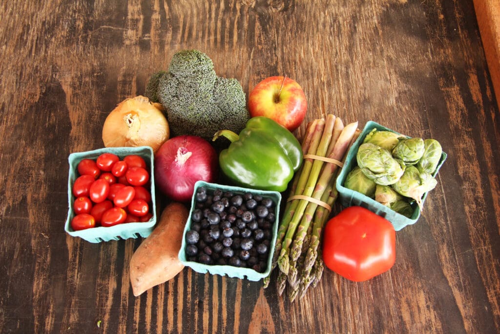 An assortment of fresh fruits and vegetables, including tomatoes, blueberries, broccoli, and asparagus, is displayed on a rustic wooden surface.