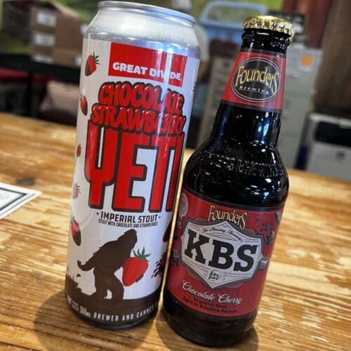 Two craft beer bottles on a wooden table: Chocolate Strawberry Yeti and Founders KBS Chocolate Cherry, placed indoors with blurred background.