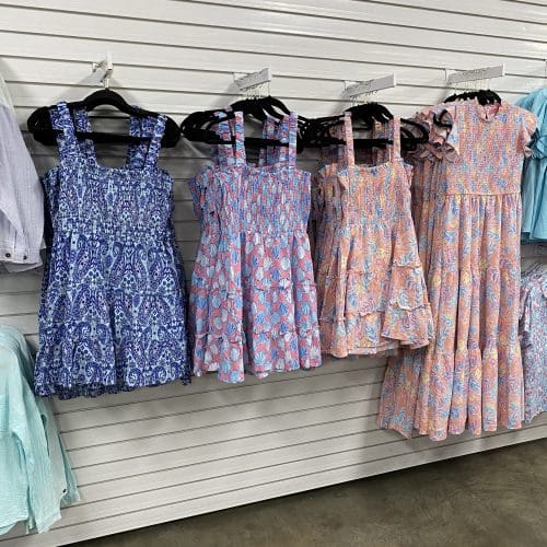 A row of colorful, patterned dresses hangs on a white slat wall in a store, alongside folded garments in a retail setting.